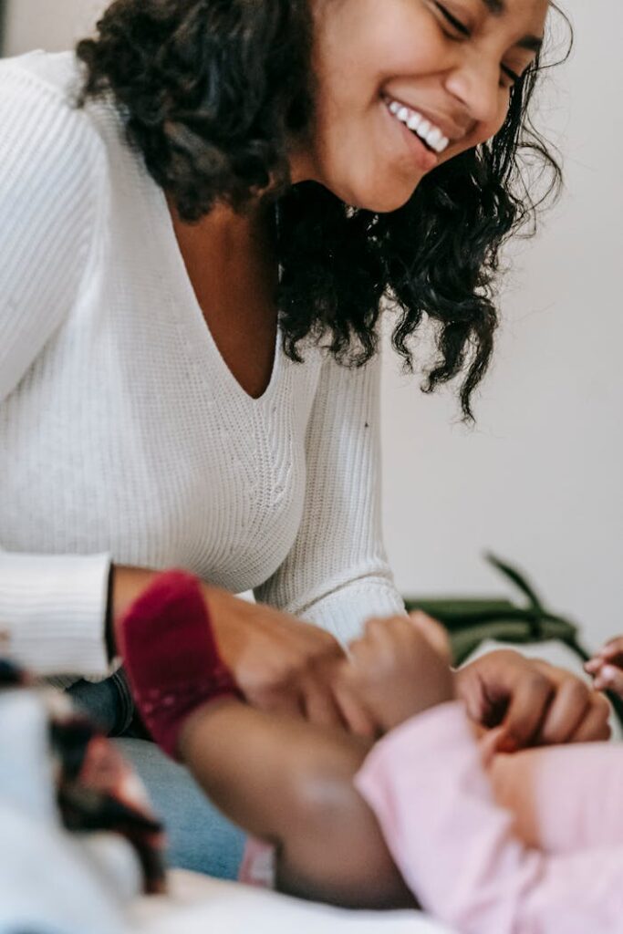 Happy African American mother smiling while putting clothes on little unrecognizable black baby lying on bed in light bedroom at home